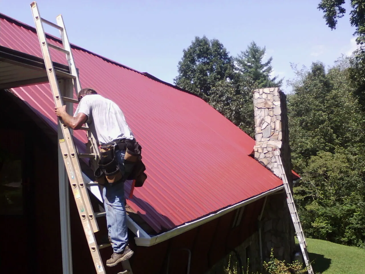 Roofer on a ladder beside a red metal roof and stone chimney — Soffit & Fascia Repair by Roofing Brothers in Gloucester City